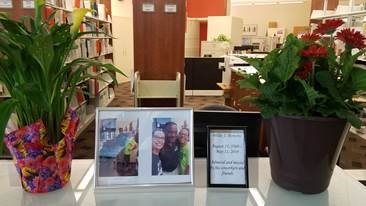 Circulation desk workspace with two flowering plants, two photos of Willie with co-workers, and a sign noting his passing